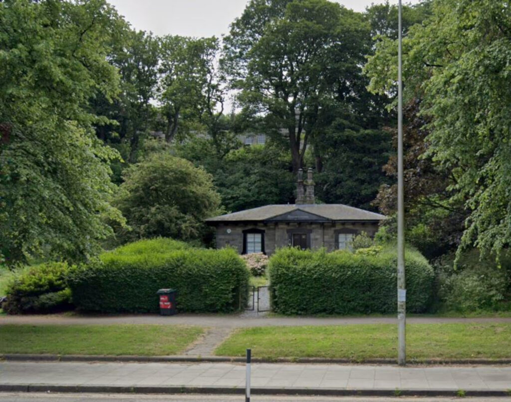 An image of a small building next to a main road, sheltered by bushes and trees.