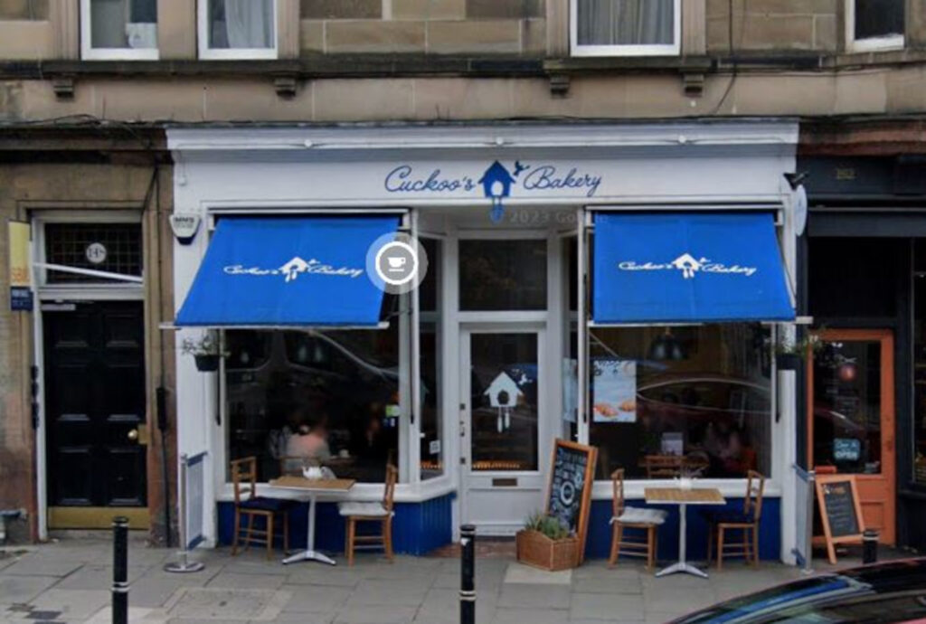 An image of a white-fronted cafe with blue awnings and tables outside on the pavement.