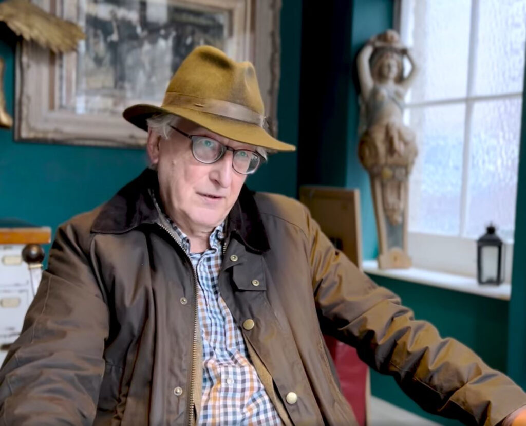 An image of an older man wearing glasses, a brown hat and brown jacket over a checked shirt.